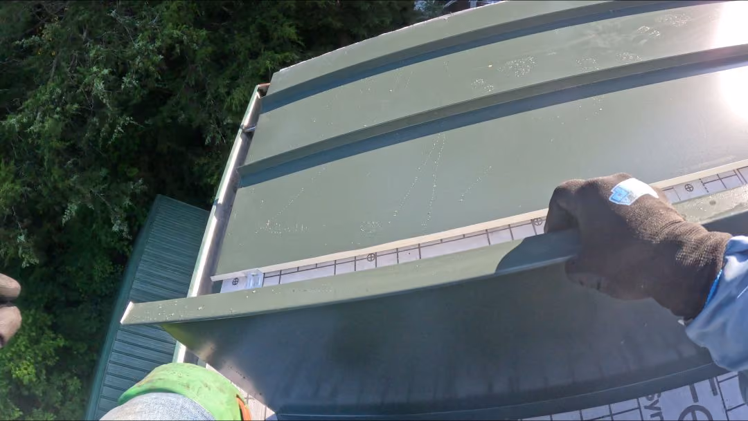 Two gloved hands installing a green metal roof panel on a roof with forest below.