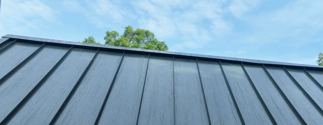 Green standing seam metal roof panels under a blue sky with trees in the background.