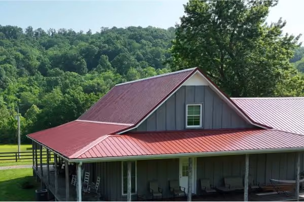 Gray house with a red metal roof surrounded by green trees and hills on a sunny day.