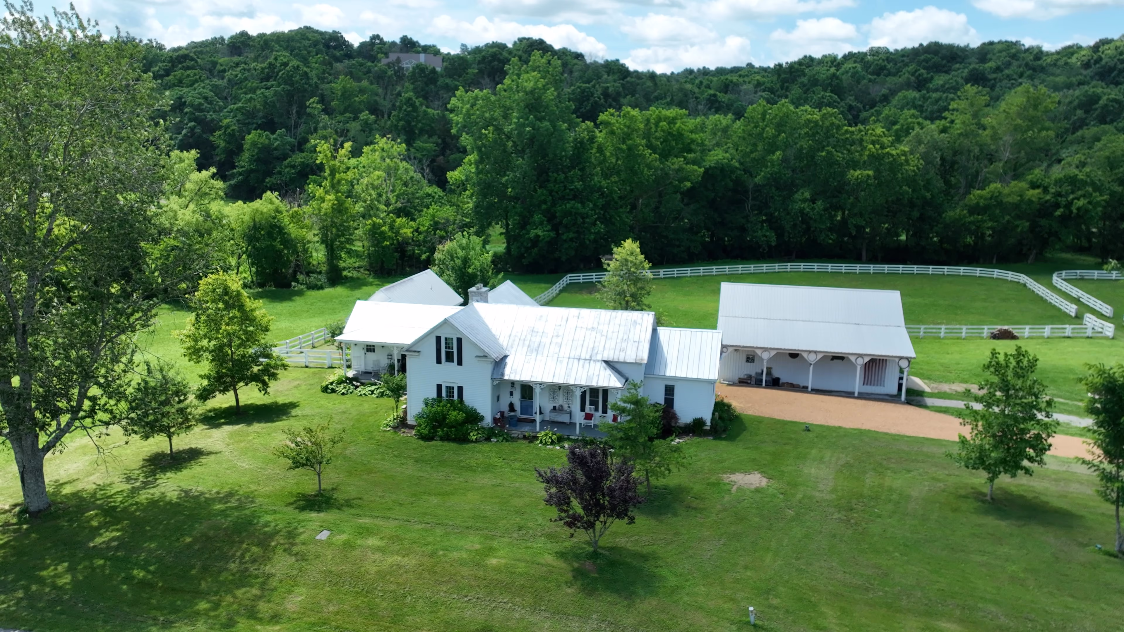 White farmhouse and barn surrounded by green trees and fenced pastures under a partly cloudy sky.
