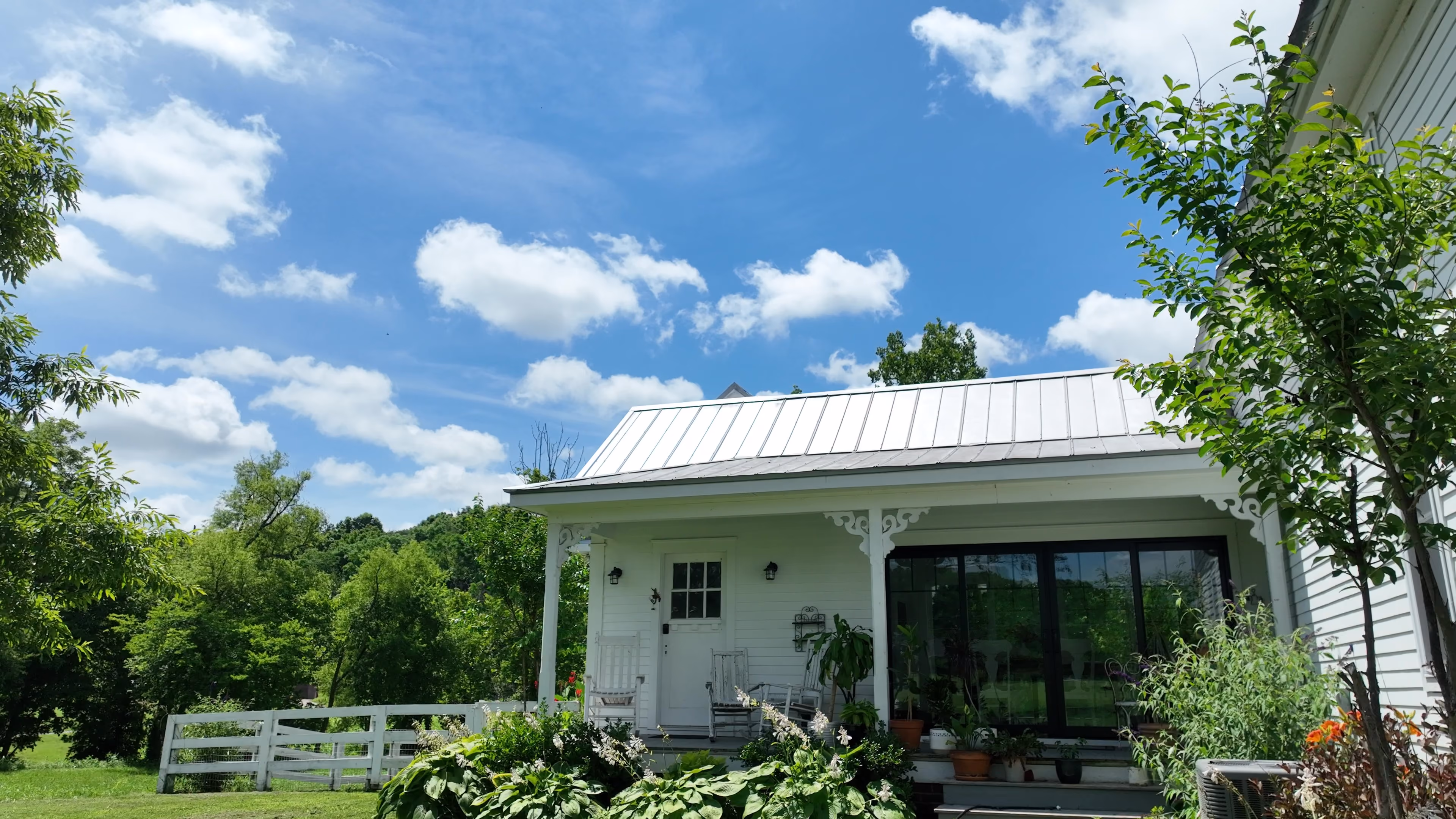 White house porch with rocking chairs and plants under blue sky with clouds amid green trees.