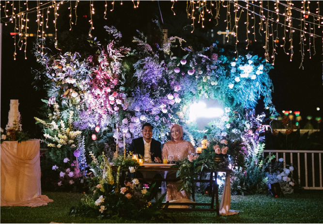 A couple smiles at their beautifully decorated table in a garden setting at night, capturing the feeling of a fairytale wedding at Whitehouse @ The Estate on Federal Hill.