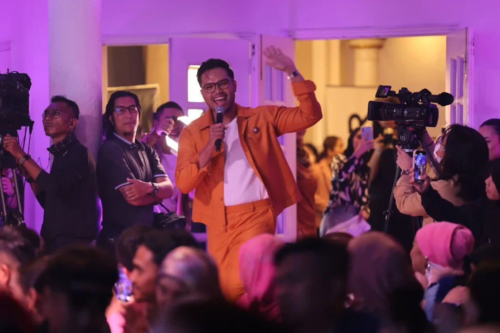 A celebrity host in a vibrant orange suit speaks on stage, highlighting the Celebrity Hosts & Guests and lively Entertainment Line-up from the launch event.