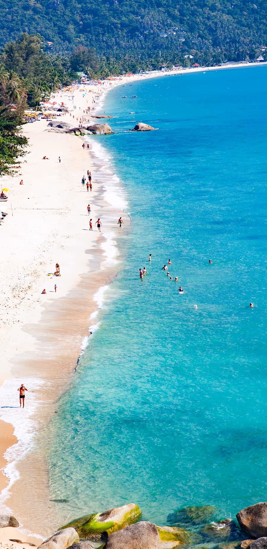 Aerial view of a tropical beach with white sand, people walking along the shore, and swimmers in clear turquoise water near lush green hills in Lamai, Koh Samui.