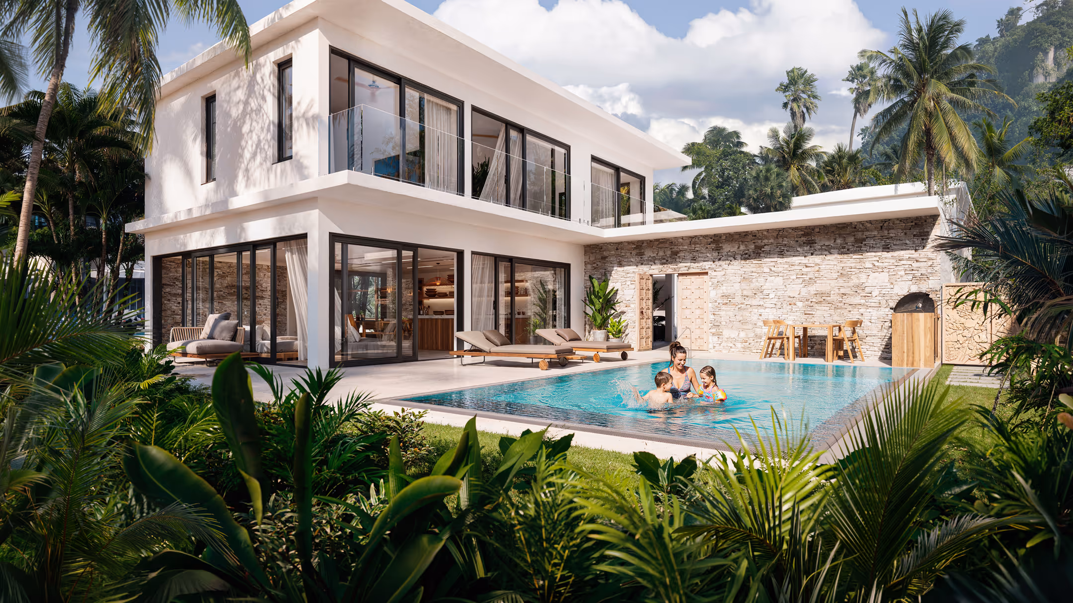 Modern two-story house with large glass windows, a swimming pool, and a woman playing with two children in the pool surrounded by tropical greenery.