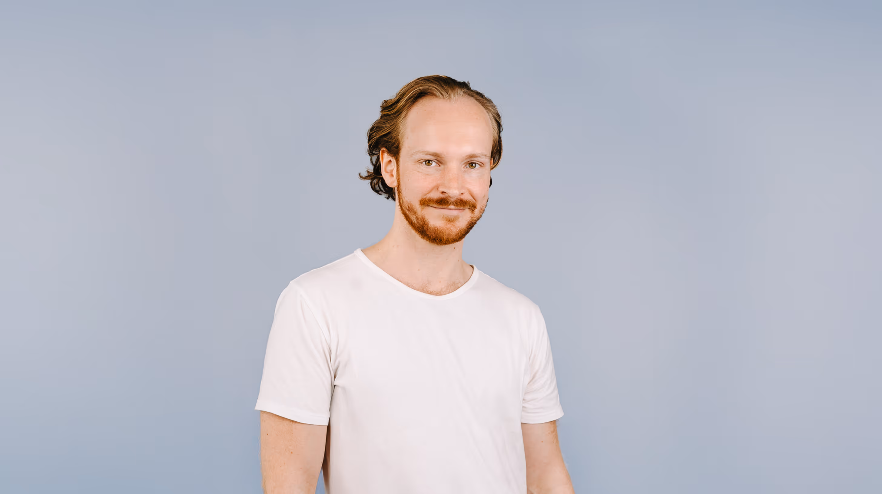 Mann mit weißem Poloshirt und grauer Hose steht lächelnd vor hellem Hintergrund mit grünen Blättern links und rechts.