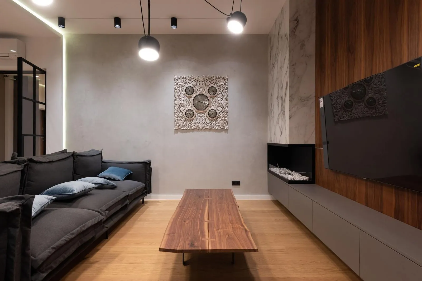 Modern living room with dark gray sofa, wooden coffee table, wall-mounted TV, and decorative wall clock with multiple time zones.