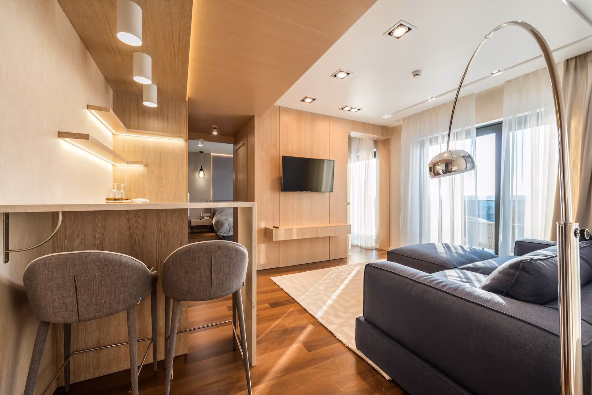 Modern open living space with a dark blue sofa, a wall-mounted TV, wooden floor, breakfast bar with two chairs, and large windows with sheer curtains.