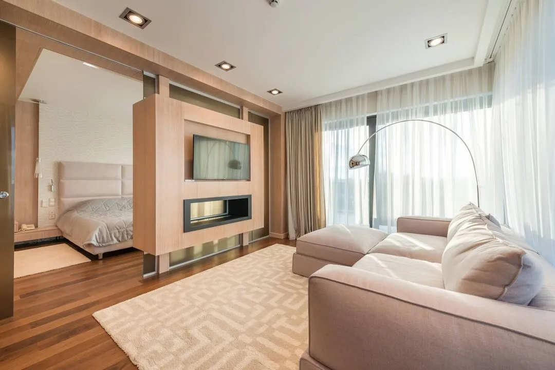 Modern living room with a beige sectional sofa, floor lamp, large window with sheer curtains, and a wooden partition wall with a mounted TV and fireplace, adjacent to a bedroom.