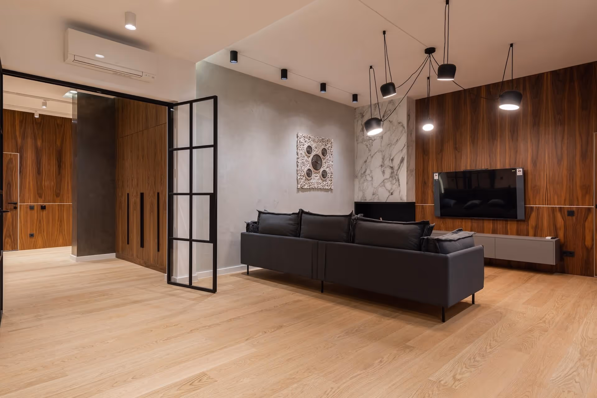 Modern living room with black sofa, wooden panel walls, wall-mounted TV, and hanging pendant lights.