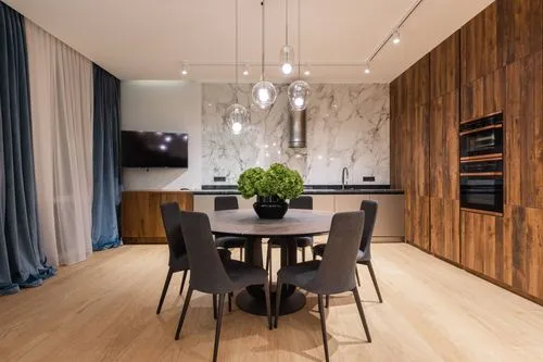 Modern dining area with round wooden table, six gray chairs, green hydrangea centerpiece, pendant lights, and kitchen with wood cabinets and marble backsplash.