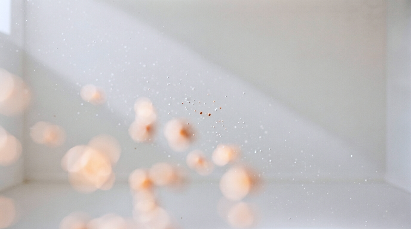 A soft shaft of daylight crossing an empty warm off-white space, revealing hundreds of tiny dust motes suspended in the air. Only two or three are in sharp focus, one of them glowing faint orange, while the rest are barely visible.