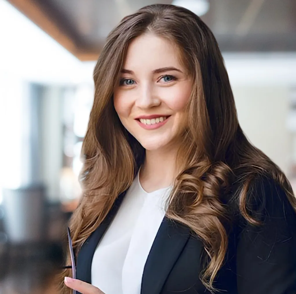 Smiling woman with long wavy brown hair wearing a black blazer and white shirt indoors.