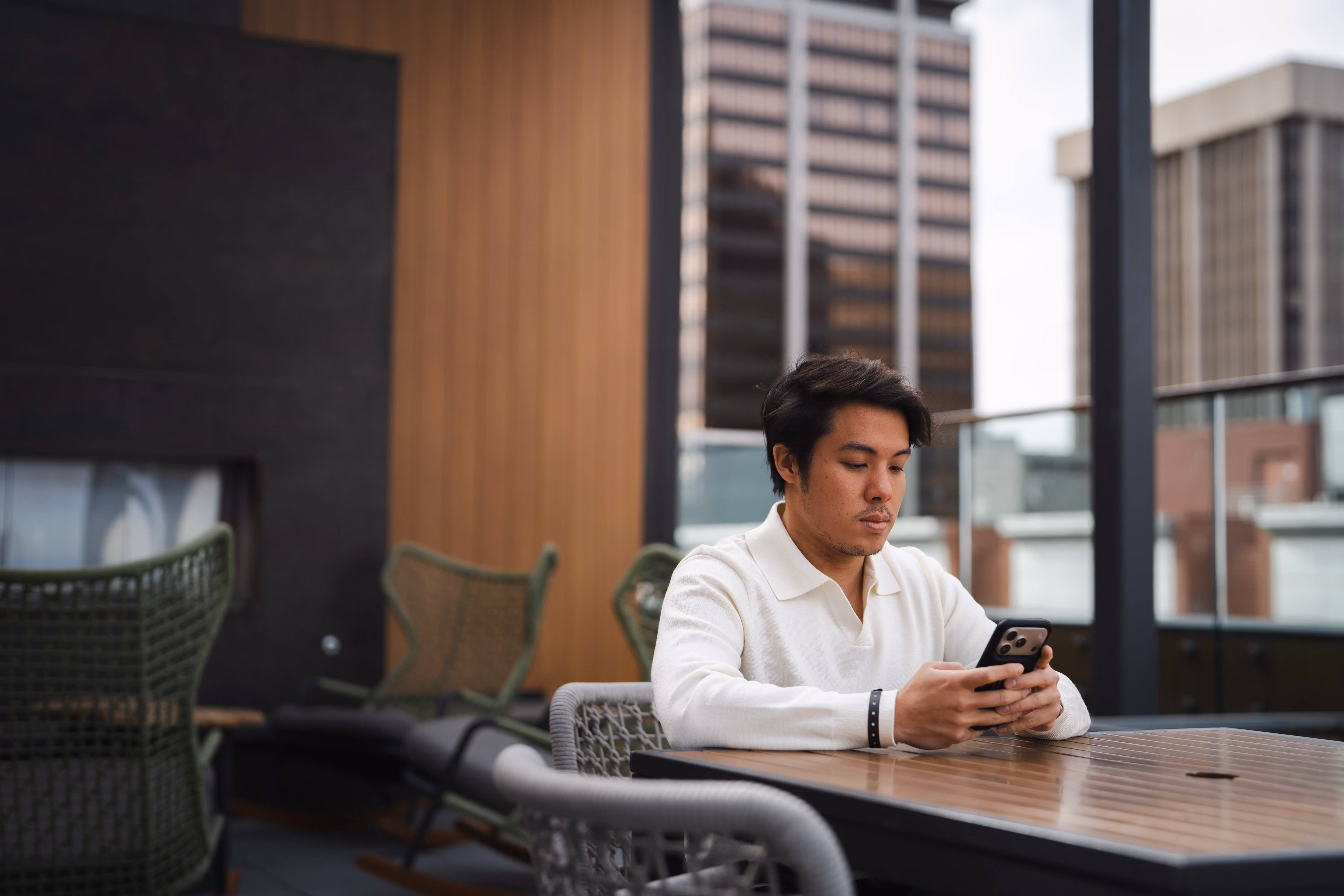 Man in white sweater sitting at outdoor table using smartphone on a rooftop patio in a city.