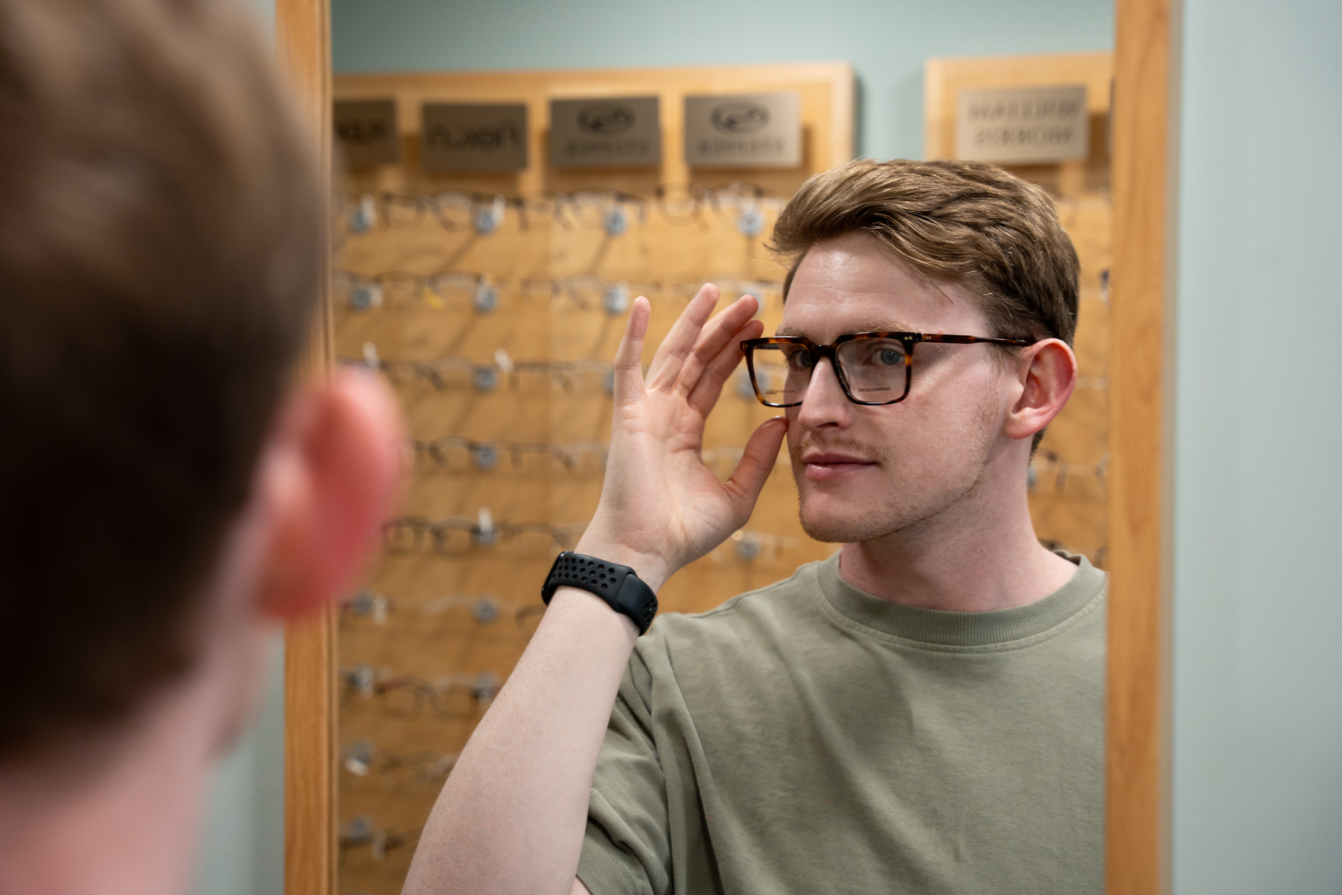A man wearing a green shirt tries on brown tortoiseshell eyeglasses in front of an optical shop mirror, adjusting the frame with his right hand, with display racks of glasses blurred in the background.