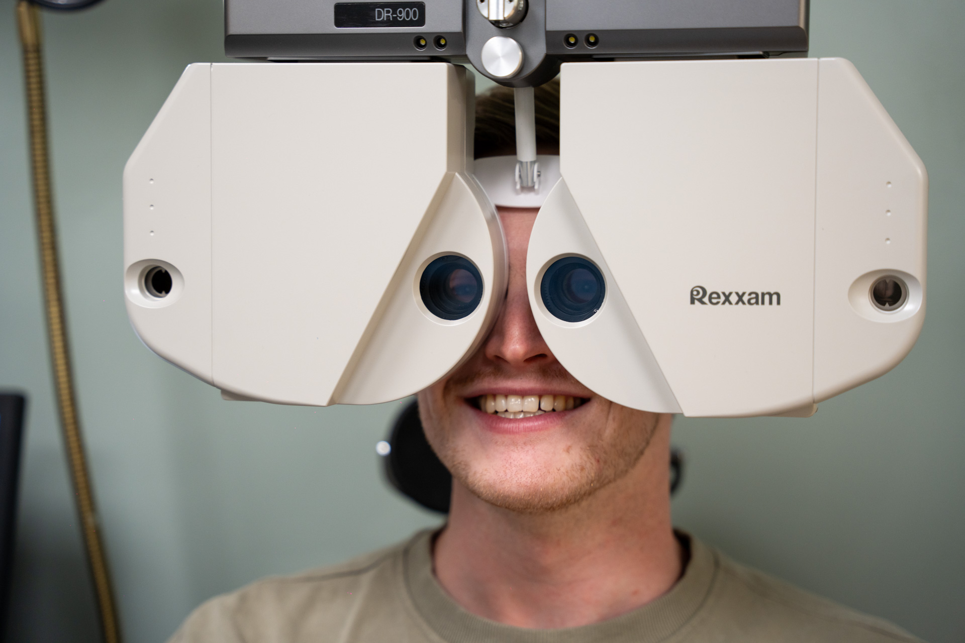 Young man smiling while having his eyes tested with a Rexxam phoropter during an eye exam.