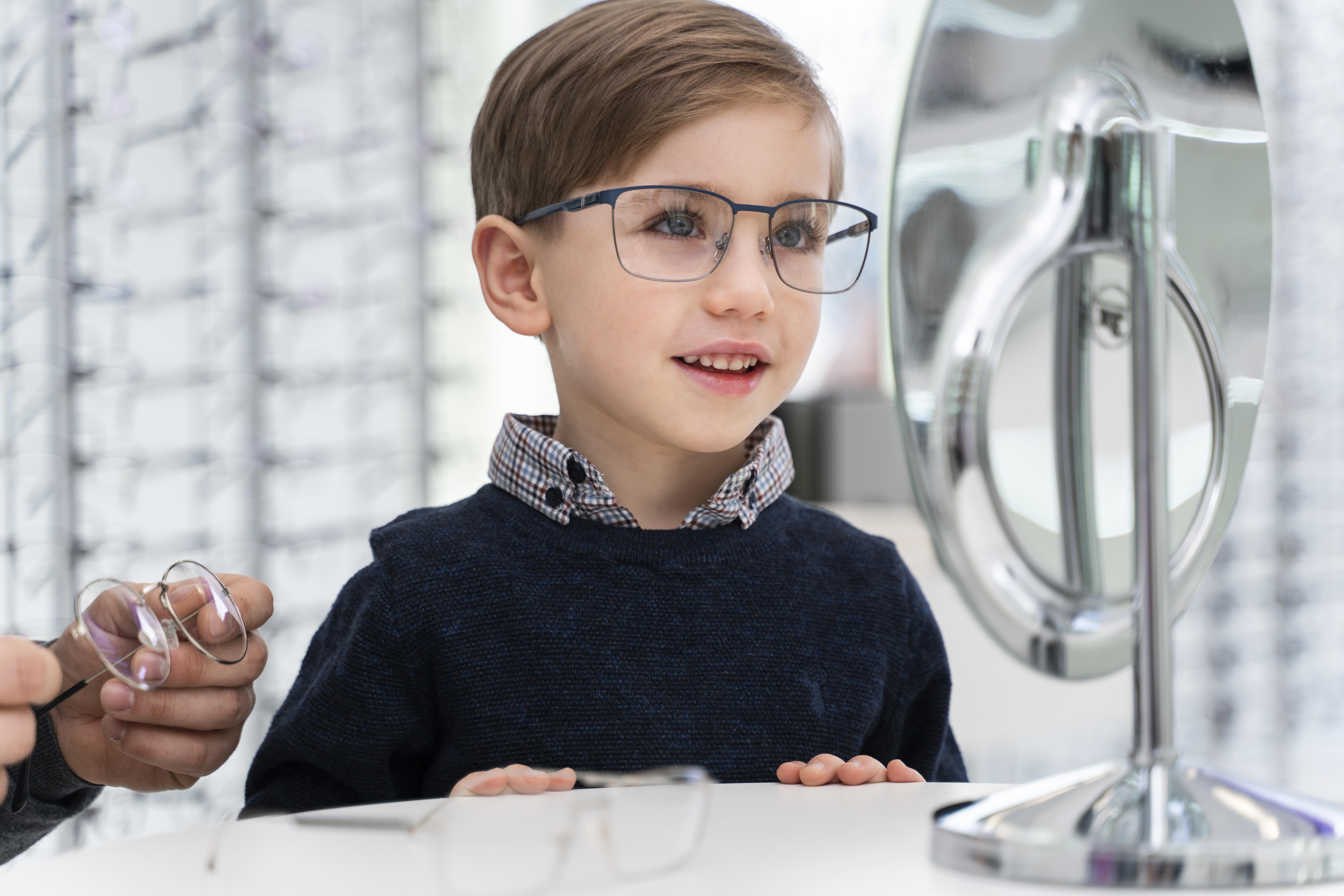 Young boy with short brown hair wearing glasses looks into a mirror at an eyewear store as an adult hand holds another pair of glasses nearby.