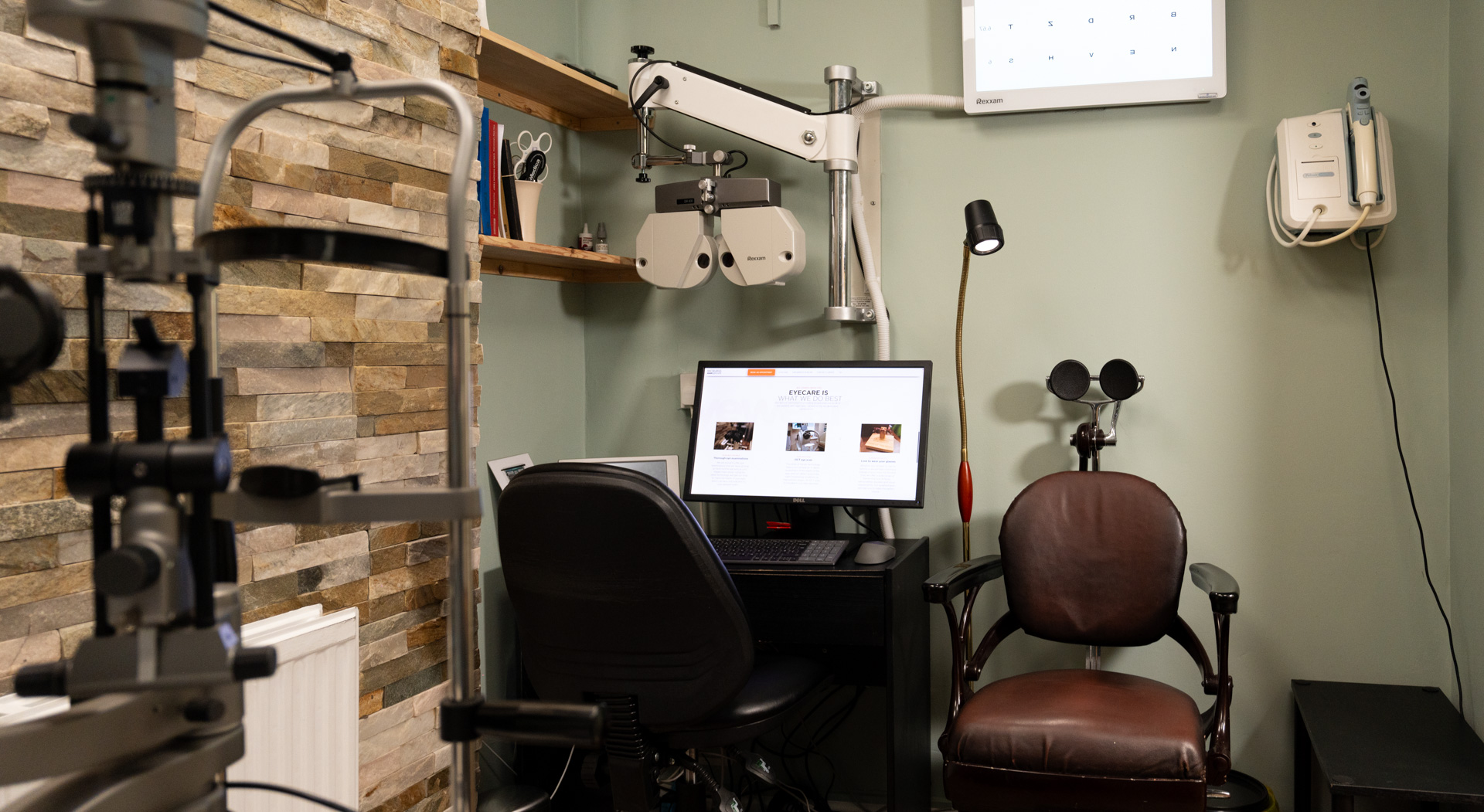 Optometrist examination room with eye testing equipment, a computer desk with chair displaying eyecare information, a brown patient chair, a wall-mounted eye chart, and shelves with books and tools.