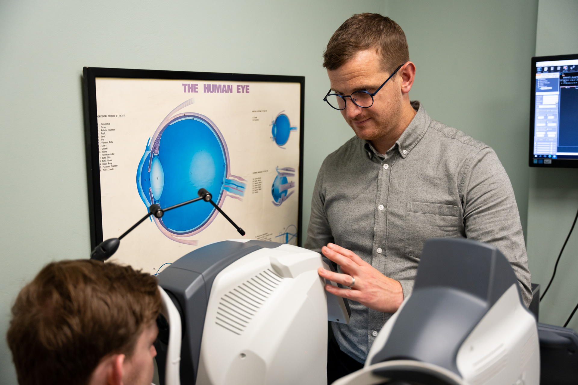 Optometrist wearing glasses and a gray shirt operating an eye examination machine while a patient rests their chin on the machine's support, with a large diagram of the human eye on the wall behind them.