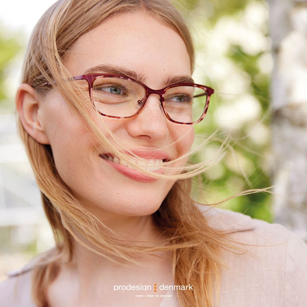 Close-up of a smiling woman wearing tortoiseshell eyeglasses with a blurred green and white outdoor background, hair gently blowing across her face.