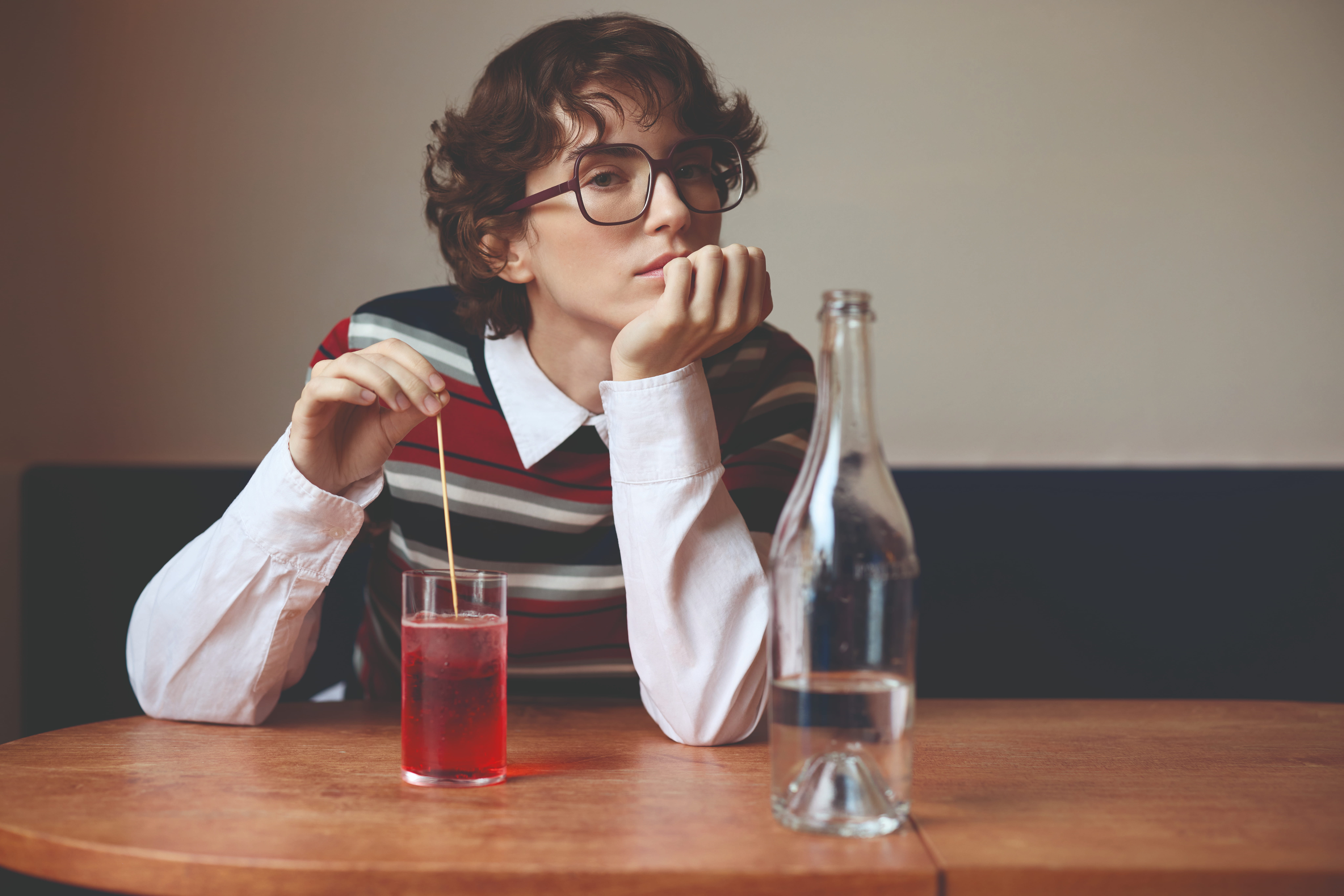 Young person with short curly hair and large glasses sitting at a wooden table, stirring a red drink with a stick, resting their chin on their other hand, with a clear bottle partially filled with water in the foreground.