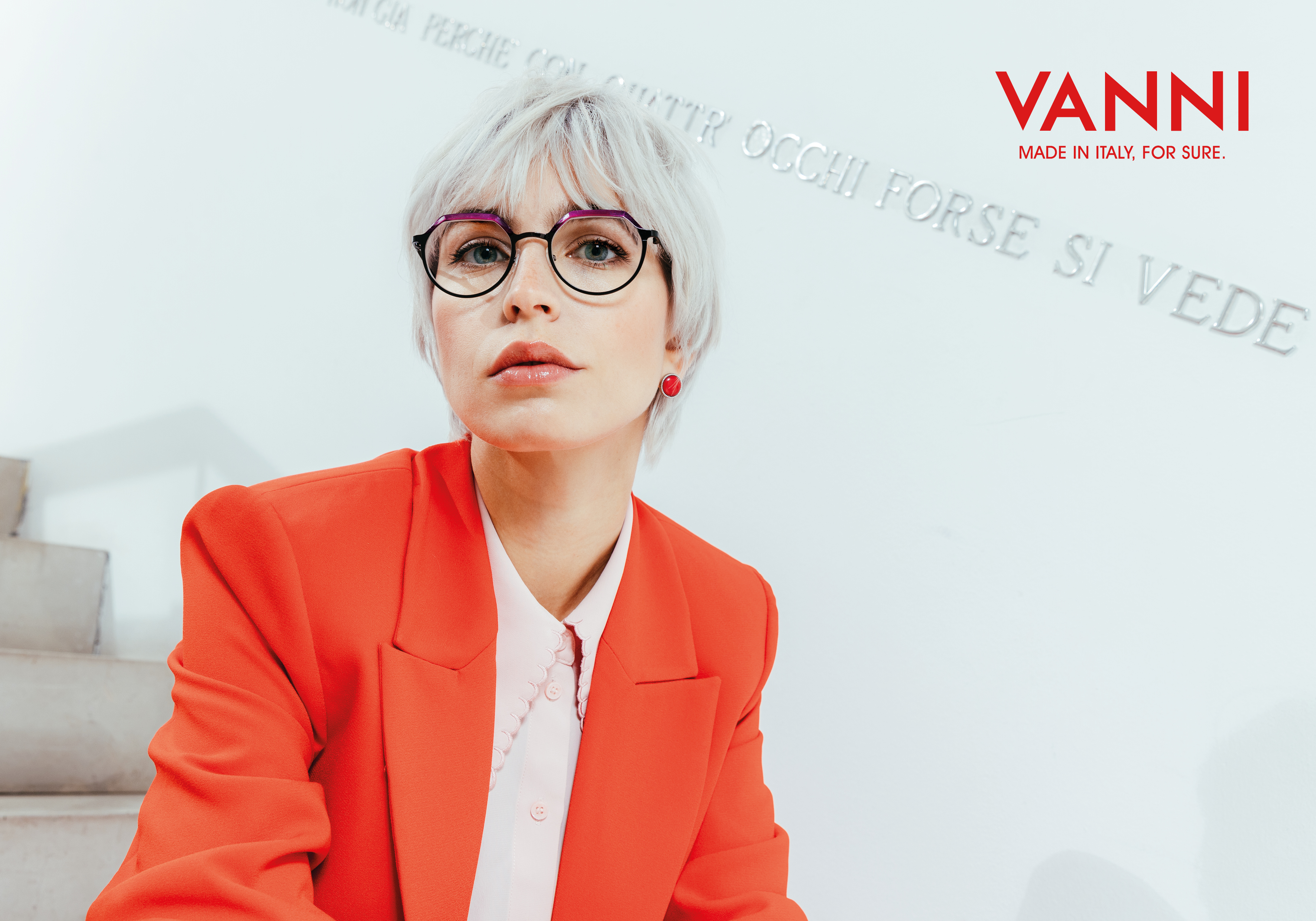 Close-up of a woman with short platinum blonde hair wearing round glasses with purple accents, red earrings, a bright red blazer, and a light pink blouse, against a white background with the VANNI logo and the text "MADE IN ITALY, FOR SURE."