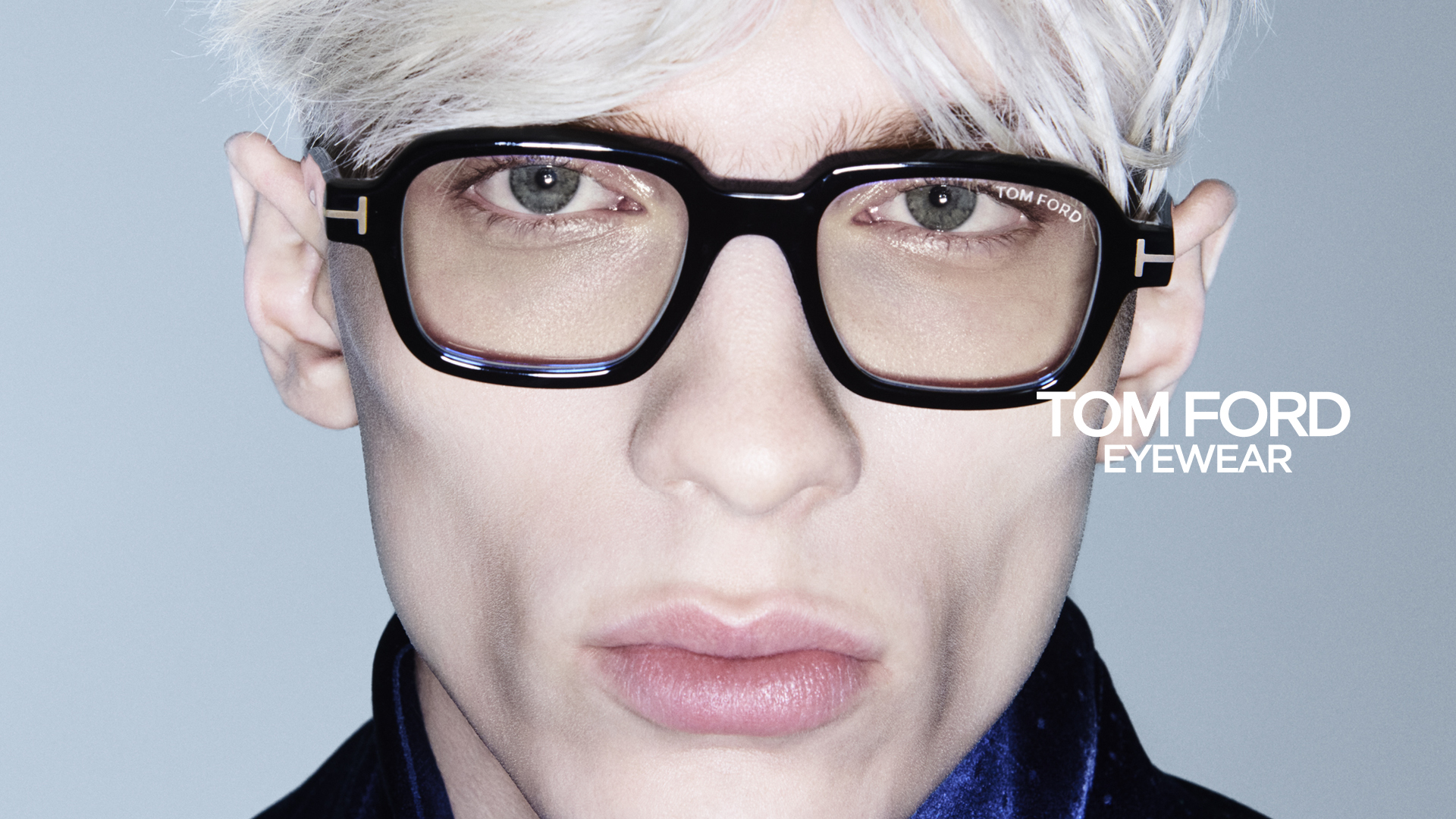Close-up of a person with platinum blonde hair wearing large black square Tom Ford eyeglasses, with text TOM FORD EYEWEAR on the right side.