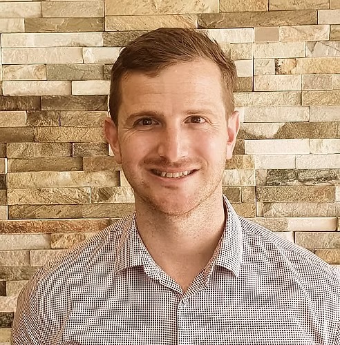Smiling man with short brown hair wearing a checkered light-colored shirt standing in front of a textured stone wall.