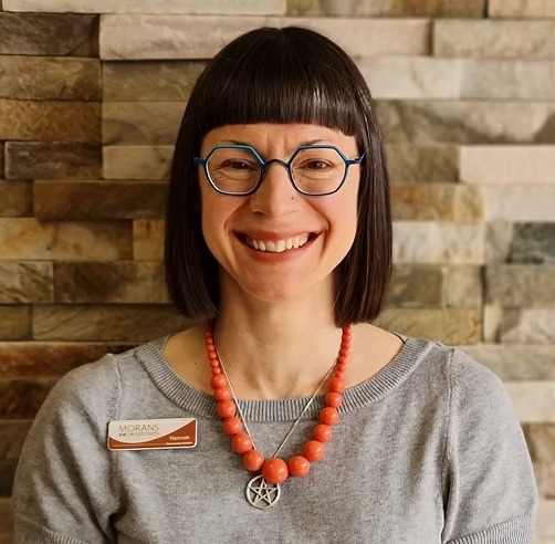 Smiling woman with short dark hair and bangs, wearing blue glasses, a gray sweater, a coral bead necklace, and a star pendant, standing in front of a stone wall.
