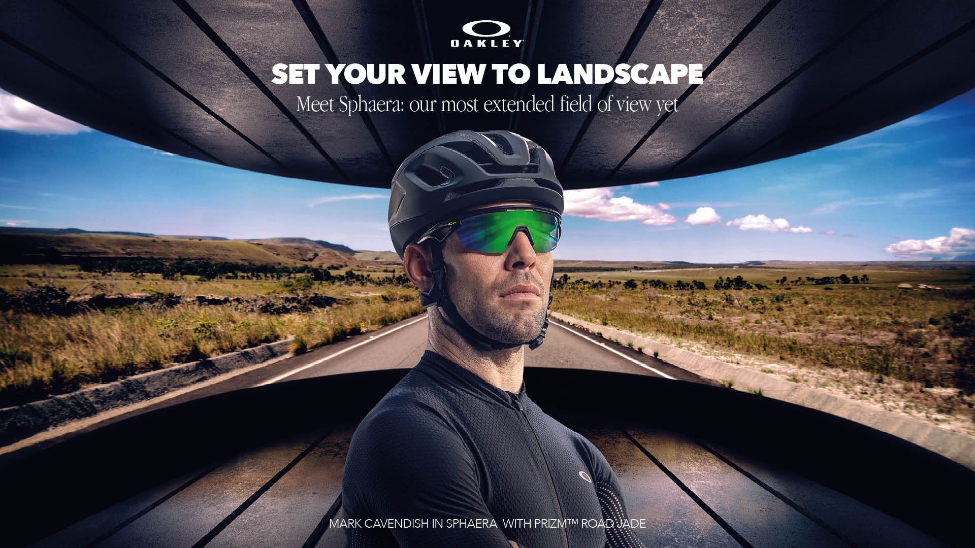 Cyclist wearing a black helmet and reflective green sunglasses stands confidently on a road with a vast landscape and blue sky behind, framed by a dark architectural structure. The text above reads 'Set your view to landscape' and mentions Oakley Sphaera sunglasses.
