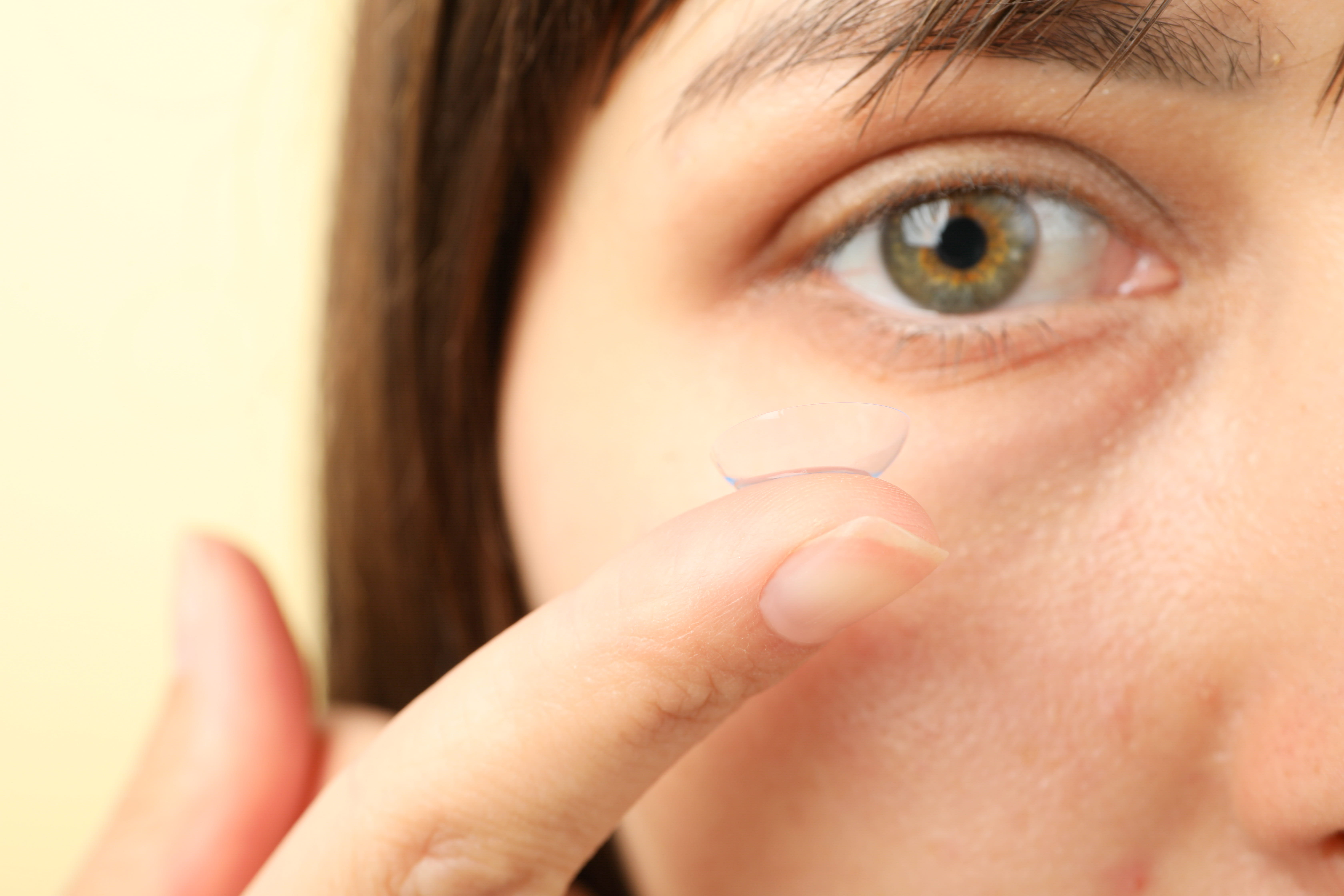 Close-up of a person's face focusing on one hazel eye and a finger holding a transparent contact lens near the eye.