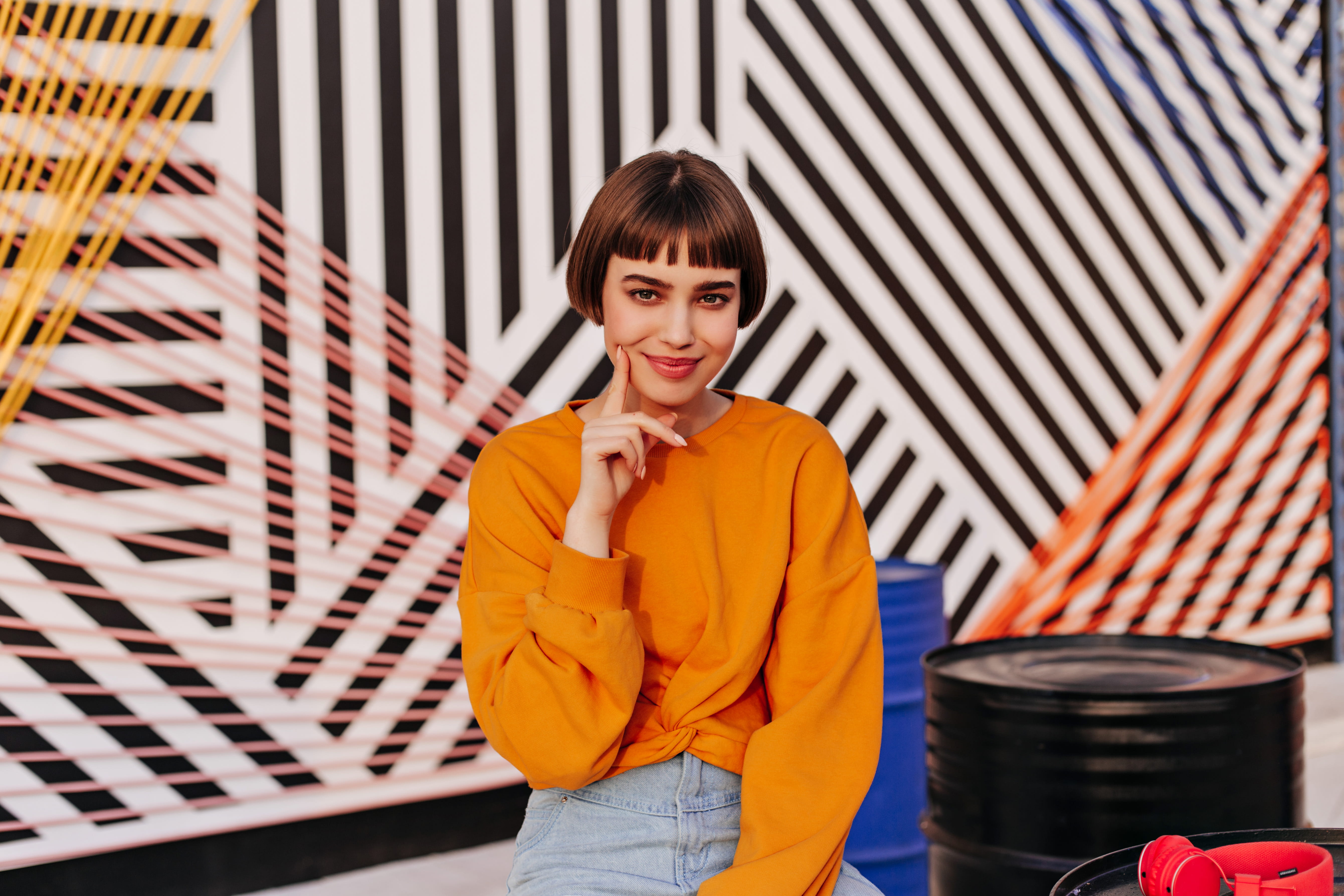 Young woman with short brown hair and bangs wearing an orange sweatshirt and light blue jeans, smiling with her finger resting on her cheek, sitting in front of a colorful geometric mural with black, white, pink, orange, and yellow diagonal lines, next to black and blue barrels and red headphones.