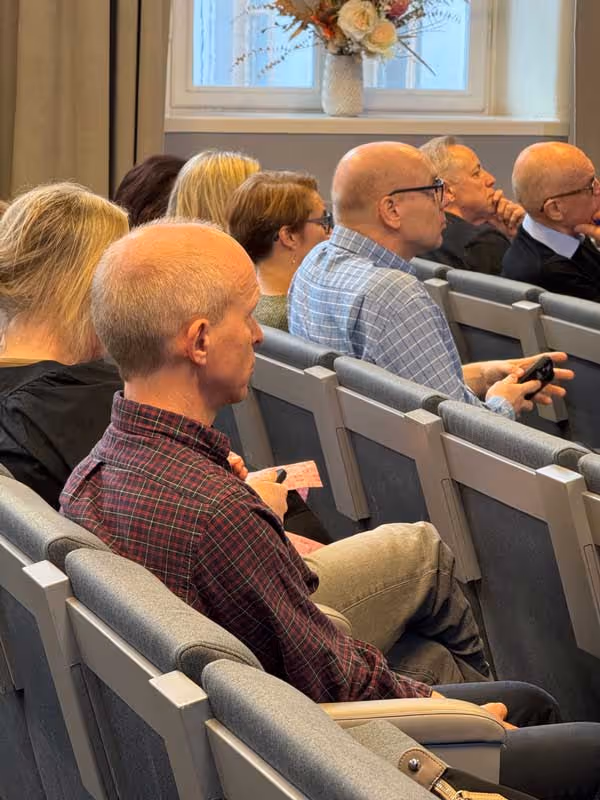 Personer sitter i stolar i ett rum och lyssnar uppmärksamt.