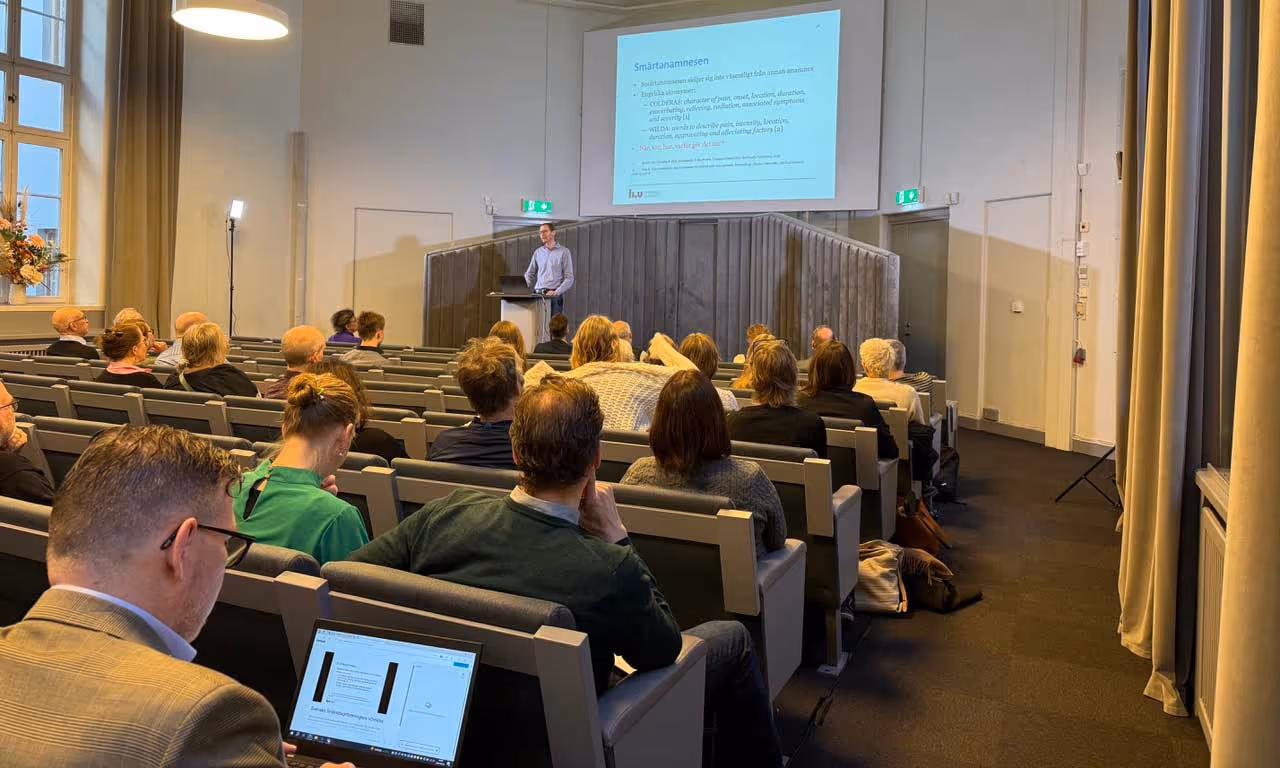 En föreläsare står vid ett podium framför en publik i en föreläsningssal med en presentationsbild projicerad på väggen.