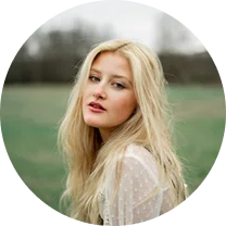 Blonde woman with long hair wearing a sheer white blouse in an outdoor setting with a blurred background.