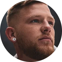Close-up of a young man with a beard, light brown hair, and a white ear gauge looking upward.