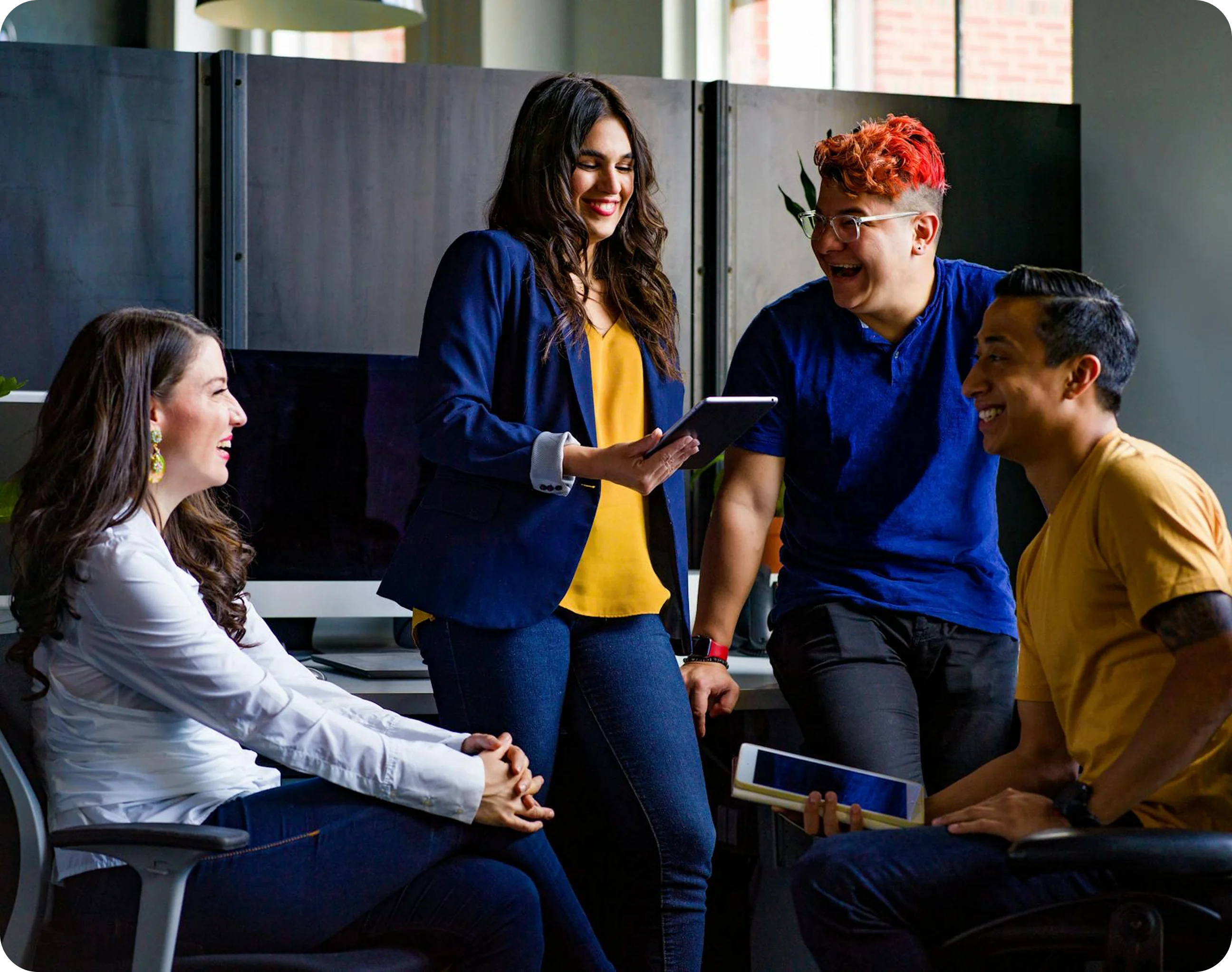 Four young adults happily discussing while holding tablets in a modern office setting.