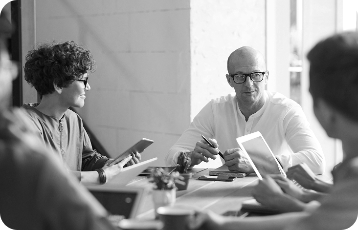 A group of four people in a meeting around a wooden table, two holding tablets, engaging in discussion in a modern office.