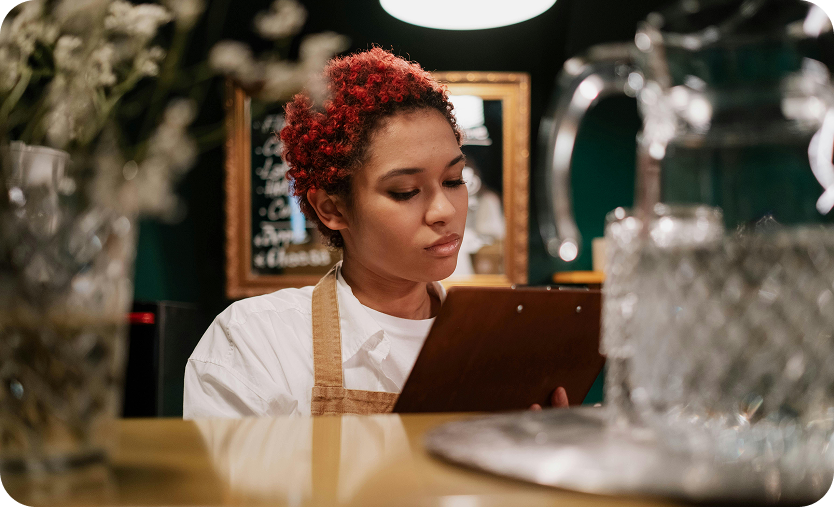 Young woman with short curly red hair wearing an apron, reading a clipboard in a cafe setting.