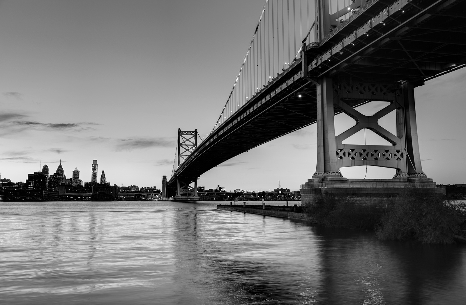 Black and white image of a large suspension bridge over a river with a city skyline in the background during dusk.