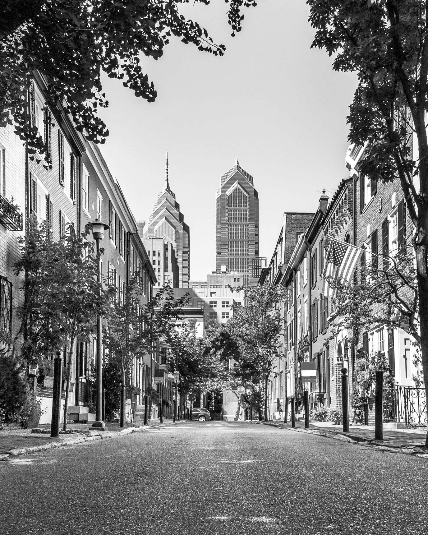 Empty urban street lined with trees and buildings, with two tall skyscrapers visible in the background under a clear sky.