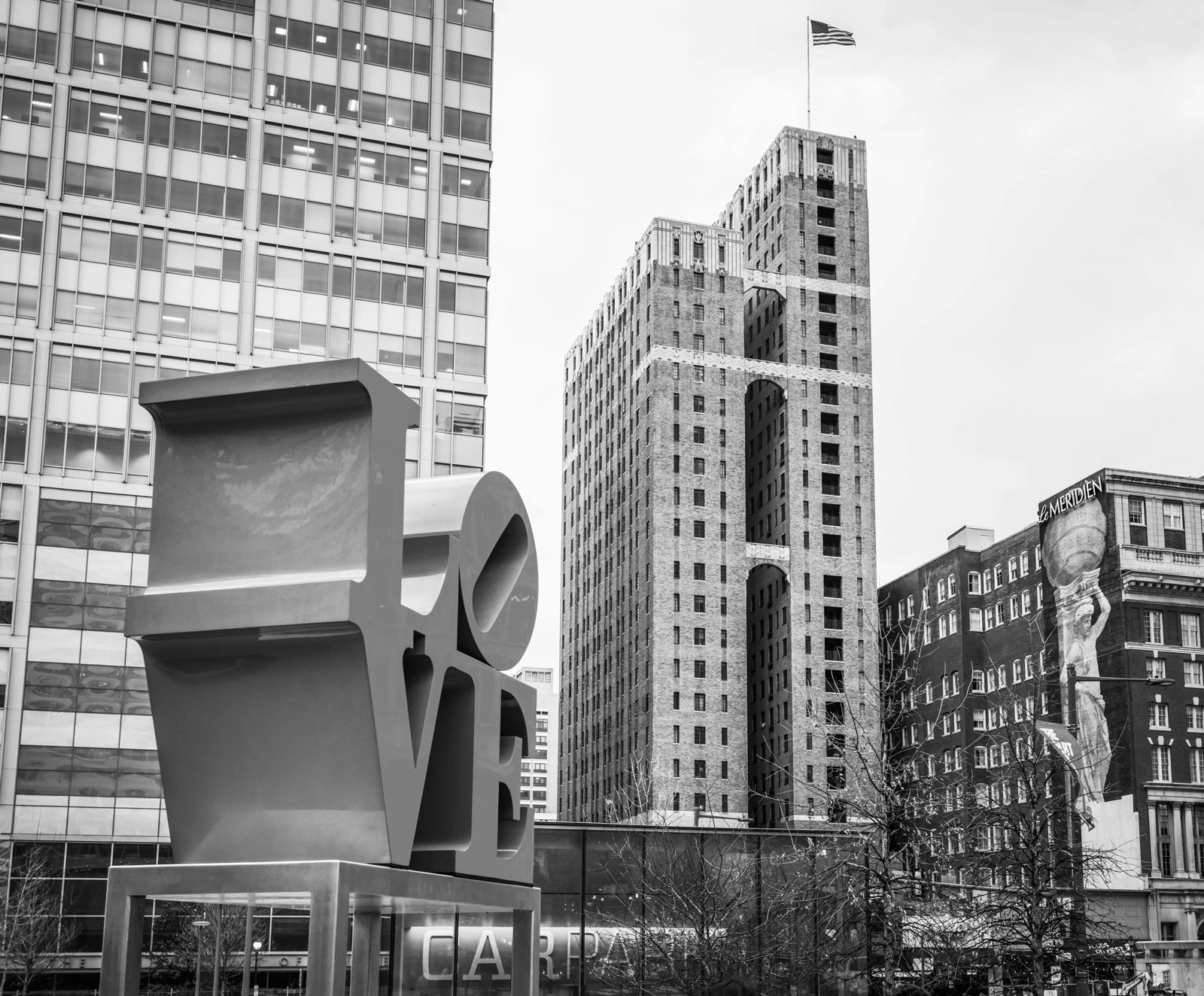 Black and white cityscape featuring a large LOVE sculpture in the foreground with tall office buildings and an American flag in the background.