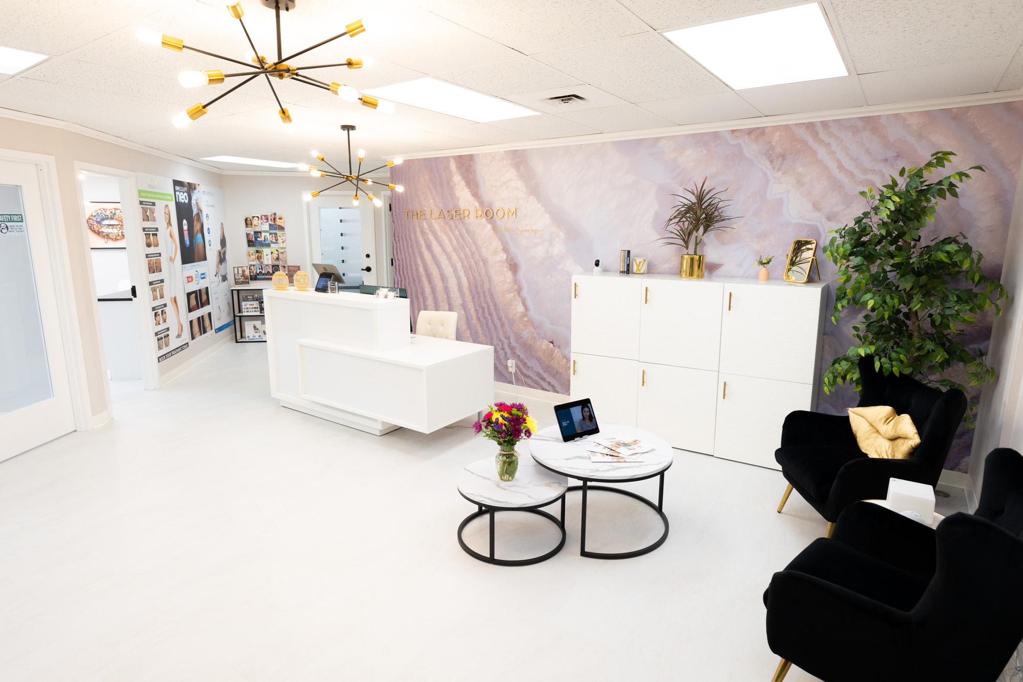 Bright modern reception area with white desk, black velvet chairs, coffee tables with flowers, a large plant, and 'The Laser Room' sign on a purple wall.