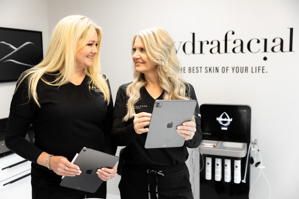 Two women in black uniforms holding iPads and smiling inside a skincare clinic with hydrafacial branding on the wall.