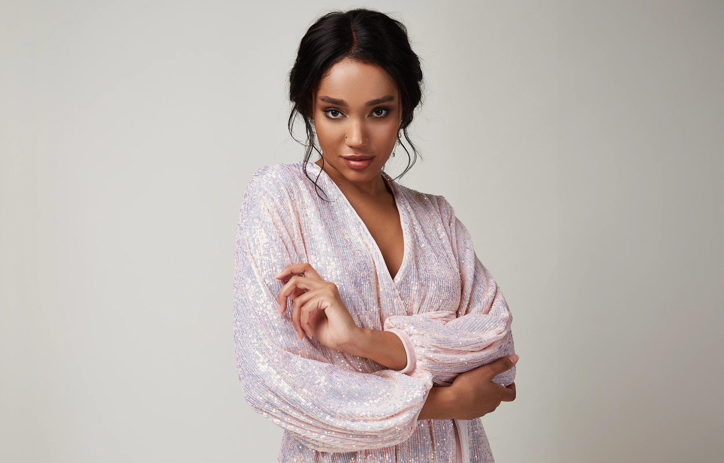 Confident woman with dark hair wearing a sparkling light pink dress posing against a plain gray background.