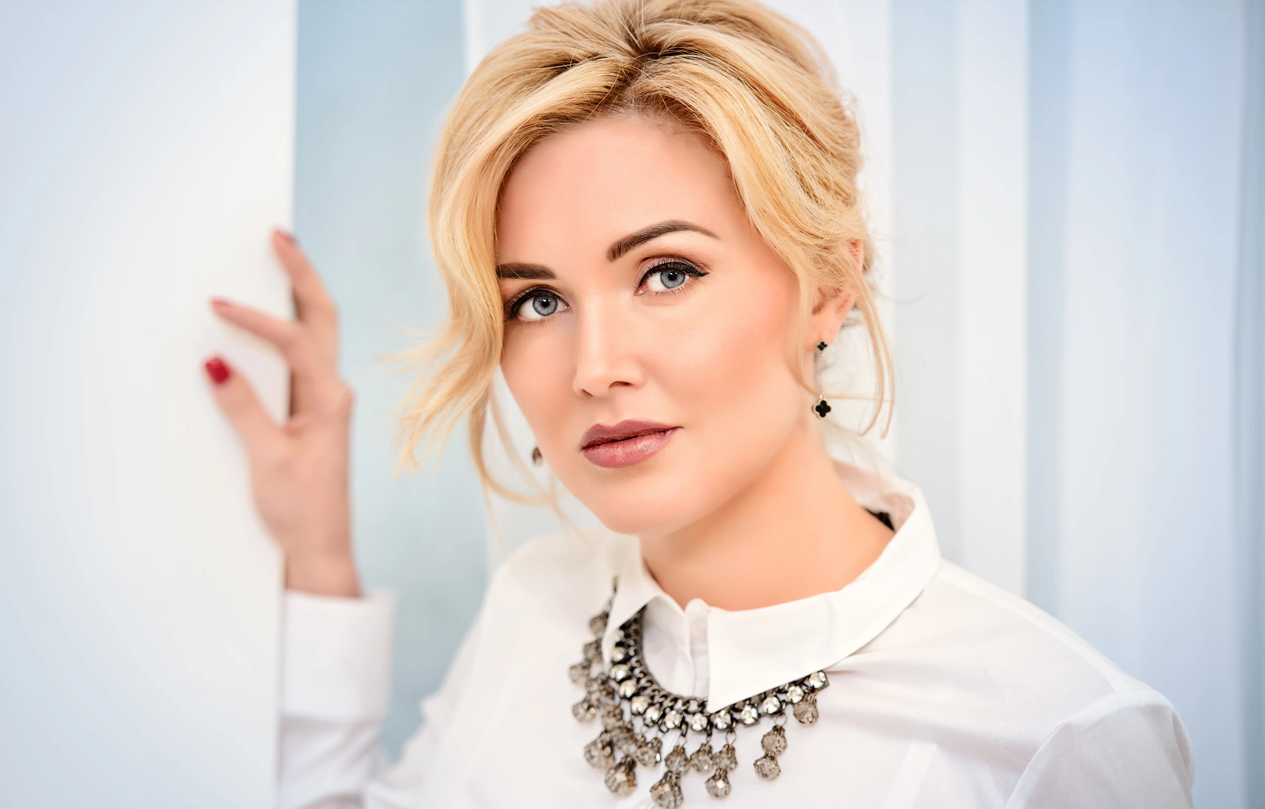 Blonde woman with light blue eyes wearing a white shirt and an ornate silver necklace, posing against a light background.