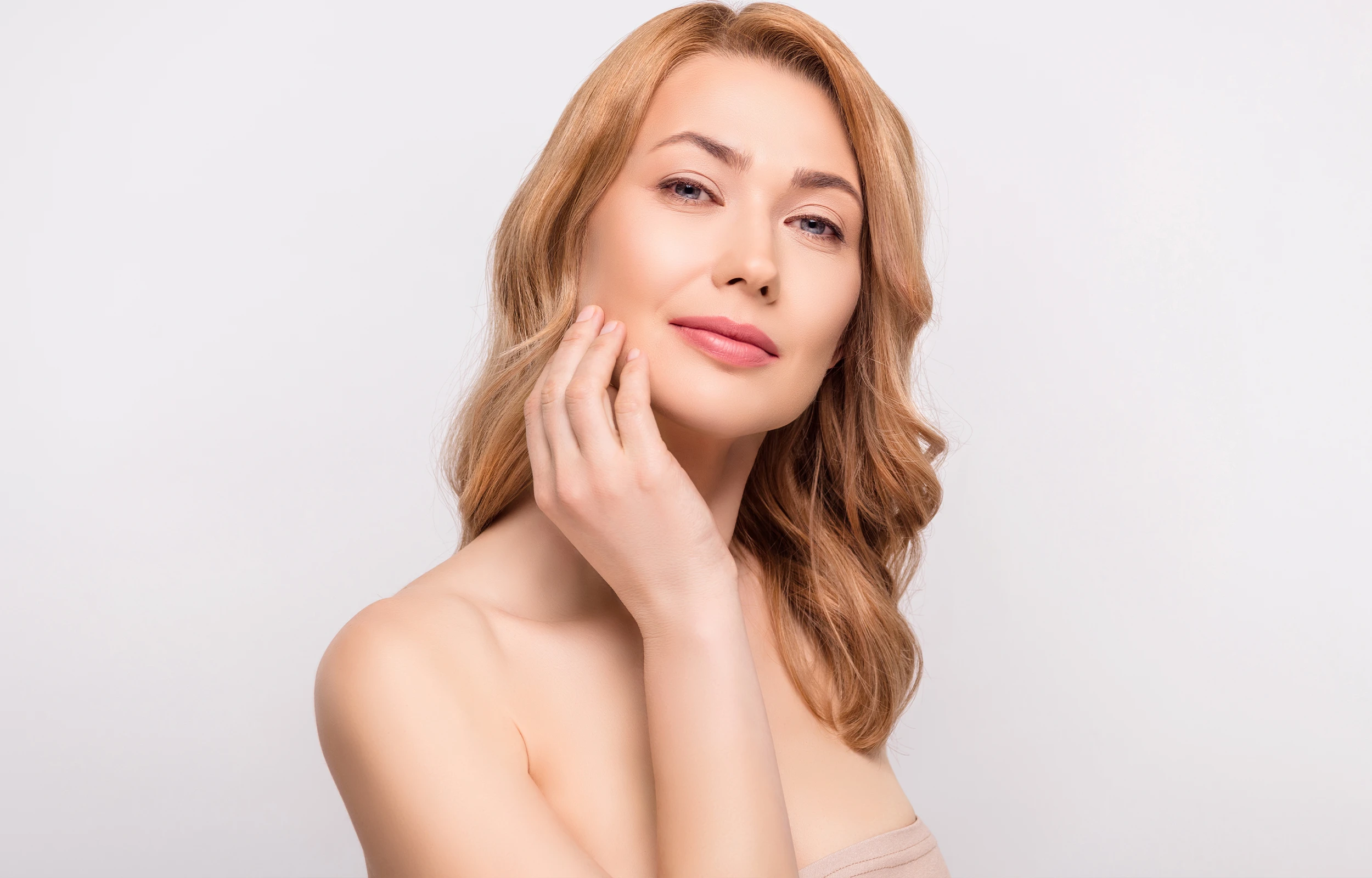 Woman with light brown hair gently touching her cheek, looking confidently at the camera.