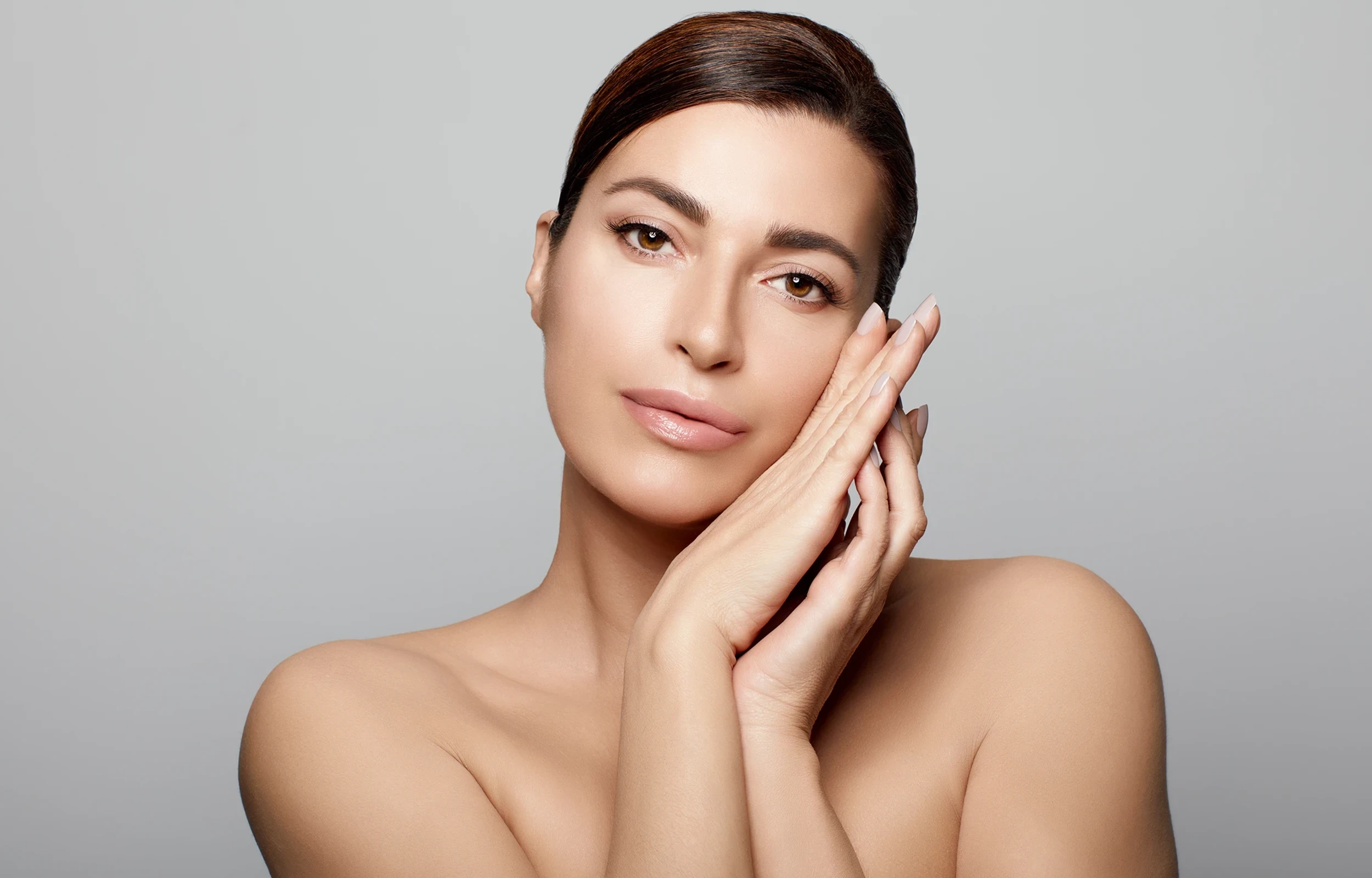 Portrait of a woman with clear skin resting her face gently on her hands against a gray background.