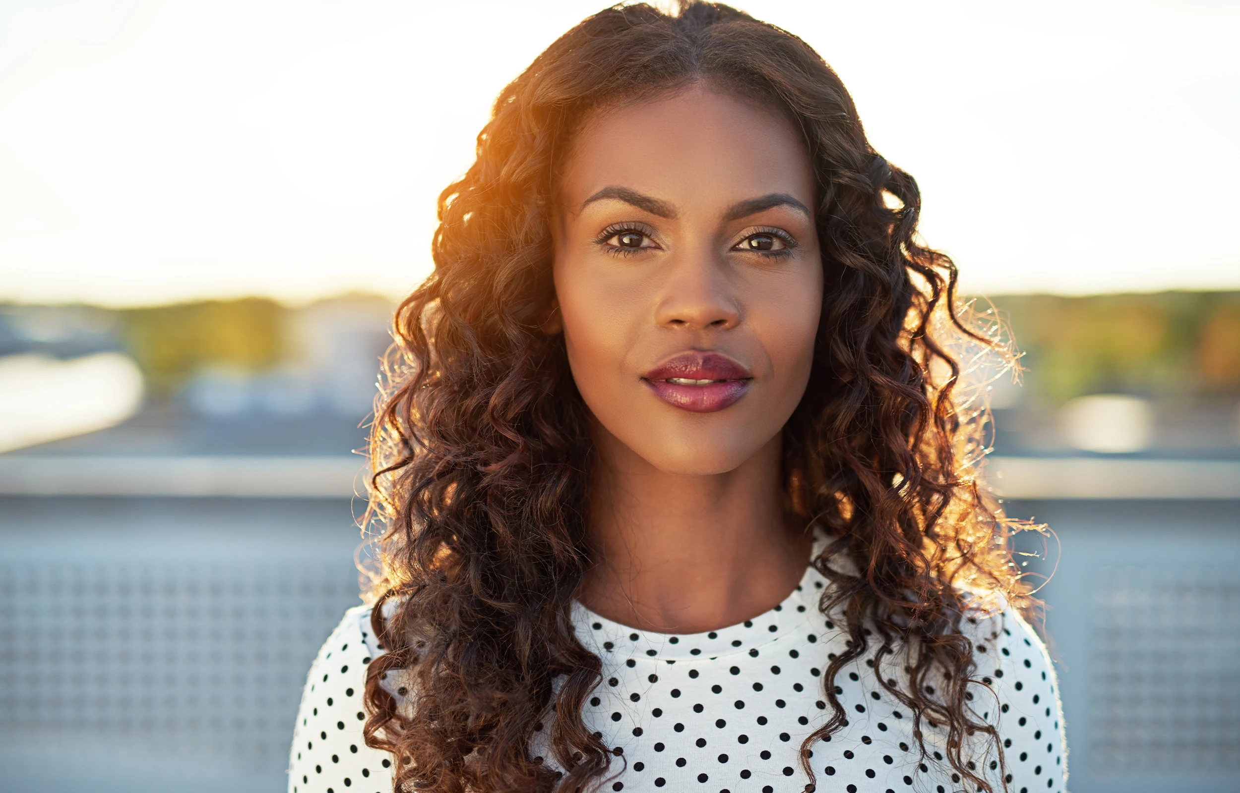 Close-up of a woman with curly hair wearing a white polka dot top, standing outdoors with sunlight behind her.