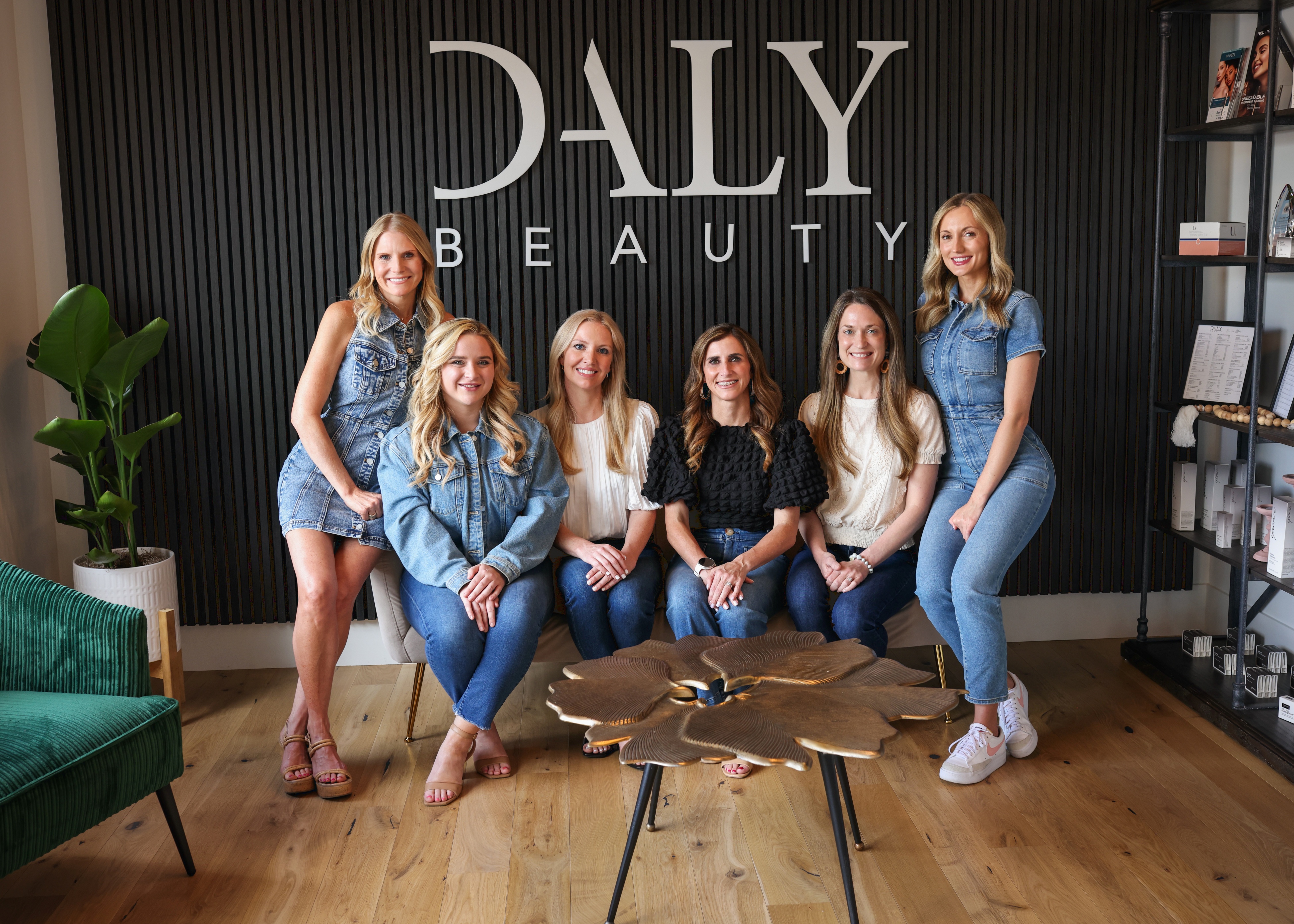 Six smiling women posing in a beauty salon with 'Daly Beauty' sign on a black slat wall behind them, seated and standing around a decorative table.